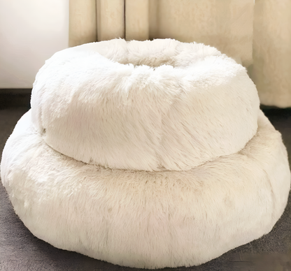 White fluffy pet bed on a dark floor with light-colored curtains in the background
